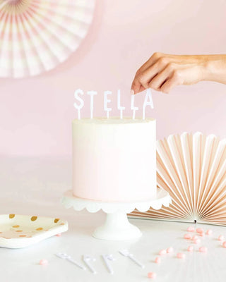 A hand places the final My Mind’s Eye Letterboard Cake Topper in white on a smooth white birthday cake by Cake by Courtney, bearing the name "stella," set against a pastel backdrop with festive decorations and candy accents.