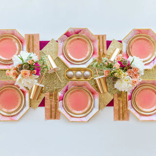 A table adorned with pink and gold plates, gold cutlery, floral centerpieces, gold cups, and a decorated runner perfectly captures the elegance of La Boheme-inspired partyware. Featuring Jollity & Co's La Boheme Cocktail Napkins, it sets an ideal scene for a summer birthday celebration.