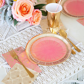 Elegant party tableware featuring pink and gold plates, gold cutlery, a gold cup, and a floral centerpiece with pink roses sits atop a woven placemat. Jollity & Co’s La Boheme Guest Napkins add to the luxurious setting for an unforgettable occasion.