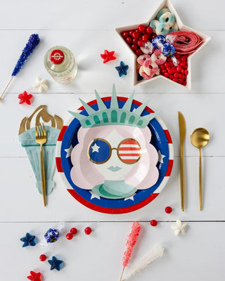 Set a festive American celebration table with Lady Liberty Red Striped Paper Plates by My Mind’s Eye, star-shaped candy bowls, Statue of Liberty plates, red and blue decorations, gold utensils, all on a white surface.