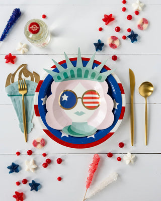 Celebrate in American style with My Mind’s Eye's Lady Liberty Red Striped Paper Plate, colorful candies, gold utensils, and a decorative torch on white—perfect for a festive table setting.