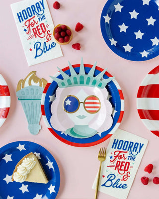 A patriotic-themed party setting featuring Lady Liberty Shaped Paper Plates by My Mind's Eye, a statue of liberty decoration, raspberries, a slice of pie, and napkins exclaiming "hooray for the".