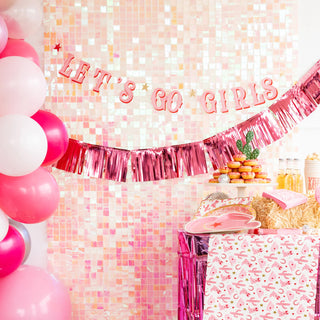 Pink-themed party decoration with a "Let's Go Girls Banner Set" by My Mind’s Eye, balloons, and gifts, featuring a sequined wall backdrop and snacks on a table.