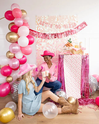 Two women celebrating with a tea party at a cowgirl-themed event, surrounded by pink balloons and festive decorations, one sitting on a stool and the other on the floor, with My Mind's Eye Pattern Paper Table Runner adorning the table.