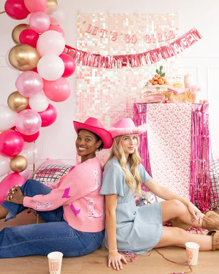 Two women in colorful outfits sitting back-to-back at a pink-themed party with balloons, a Let's Go Girls Banner Set by My Mind's Eye, and party decorations.
