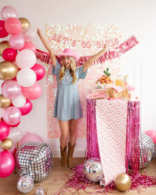 Woman in a blue dress and cowboy boots celebrating, standing between pink and gold balloons and a table with snacks, under a "Let's Go Girls Banner Set" sign by My Mind’s Eye.
