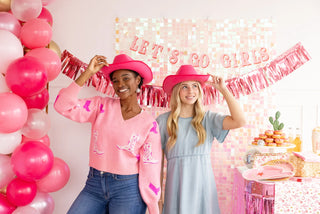 Two women in pink hats smiling at a party with pink balloons and a My Mind’s Eye Let's Go Girls Banner Set in the background.
