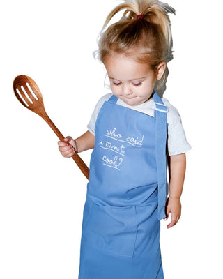 A young child wearing a Pop! Party Supplies Linen Cooking Aprons that says "who said I can't cook?" holds a wooden spoon and looks downward.