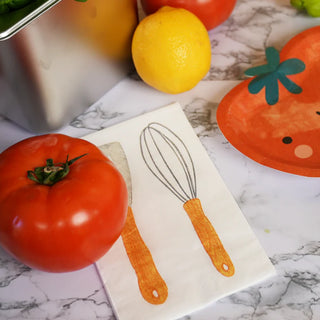 A tomato and a lemon on a marble countertop next to Little Chef Napkins by Pop! Party Supplies, decorated with a cooking utensil design, featuring a whisk and knife illustration.