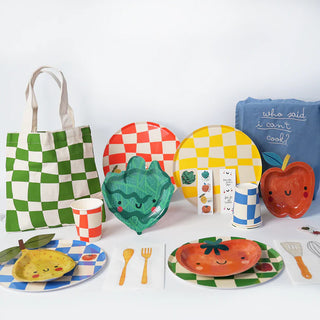 An assortment of colorful, fruit-themed tableware items including plates, sustainable FSC paper napkins, and Pop! Party Supplies Little Chef Paper Party Cups arranged with a green checkered design tote bag and a blue apron with text that reads "who said I can't cook!".