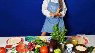 Person wearing a blue apron stands behind a table with assorted vegetables, herbs, and two empty pizza bases in a kitchen setting, surrounded by Pop! Party Supplies Little Chef Paper Party Cups.