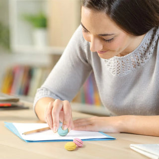 A woman in a gray sweater sits at a desk, using a blue Fred and Friends Macaron Eraser as a stamp on paper. Two strawberry-scented Fred and Friends Macaron Erasers rest in front of her, with colorful books in the background.