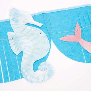 The Mermaid Fringe Garland by Meri Meri, featuring paper decorations with a pastel blue seahorse and pink mermaid tail hanging on a string against a blue fringed backdrop, adds the perfect touch to an under-the-sea party.