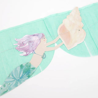 Illustration of a mermaid with purple hair holding a large seashell against a teal background, perfect for an under-the-sea party, featured on the Mermaid Fringe Garland by Meri Meri.