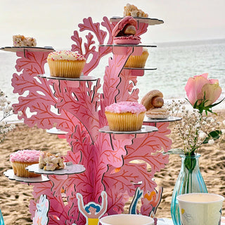 A delightful beach-themed children's party features the Talking Tables Mermaid Party Cake Stand adorned with tiers of assorted cupcakes and pastries. Small vases filled with flowers surround the pink decorative stand, perfectly embracing a plastic-free theme.