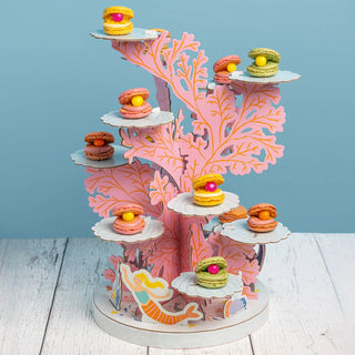 The Mermaid Party Cake Stand from Talking Tables is a coral reef-themed, plastic-free stand that displays colorful macarons on seashell plates, featuring a mermaid illustration at the base against a light blue background—perfect for any children's party.