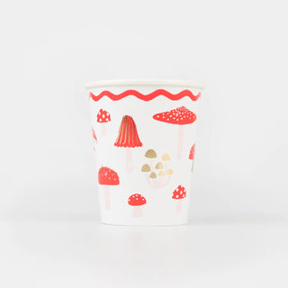 A Merry Mushrooms Cup by Meri Meri, featuring a white base adorned with red and beige mushroom illustrations, gold foil accents, and a red wavy line near the rim, stands on a white surface against a plain background.