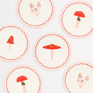 Illustration of six whimsical Merry Mushrooms Side Plates by Meri Meri, each showcasing different types of mushrooms on a beige background with red decorative borders and gold foil details.