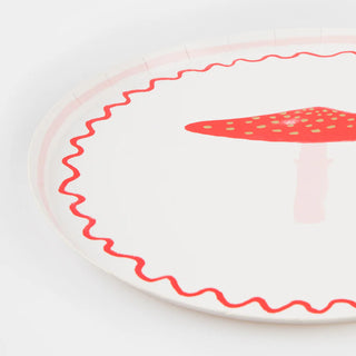 The Merry Mushrooms Side Plates by Meri Meri are round paper plates with a red mushroom illustration at the center, surrounded by red wavy lines near the edges. The festive design features small paper plates accented with gold foil details for added charm.