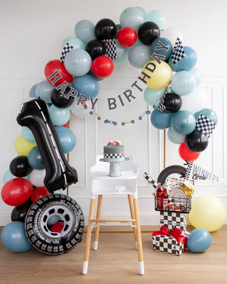 A birthday celebration with racing-themed decorations, including a My Mind’s Eye Miles Per Hour - Happy Birthday Banner Set, colorful cars, a balloon arch, a large inflatable tire and number one, a small cake on a high chair, and a cart with checkered flags and snacks.