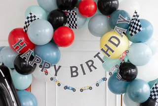 A "Miles Per Hour - Happy Birthday Banner Set" by My Mind’s Eye is surrounded by a festive arrangement of multicolor balloons and checkered flags, featuring vibrant illustrations of colorful cars. This lively party accessory is perfect for celebrating the special day with a racing theme.