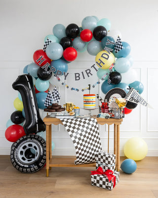 A birthday party setup with a racing theme. It features a balloon arch, a colorful My Mind’s Eye Miles Per Hour - Happy Birthday Banner Set, a cake, presents wrapped in checkered paper, and various party decorations on a table.