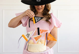 An individual donning a large black hat and a pink dress places a Halloween Mini Felt Pennant Banner from My Mind's Eye, featuring the word "EEK!", atop a festively decorated cake. The cake, embellished with vibrant Halloween decorations and phrases such as "BOO!" and "Spooky," is ideal for any spooky celebration.