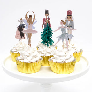 The Nutcracker Cupcake Kit by Meri Meri features Christmas cupcakes topped with paper figures of dancers and characters, beautifully arranged on a white cake stand in shimmering gold foil cases.