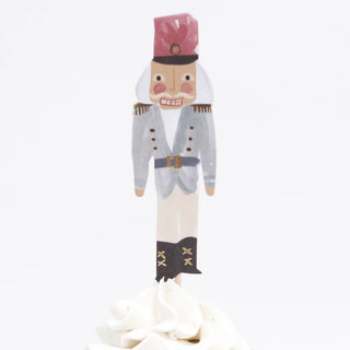 A Nutcracker Cupcake Kit by Meri Meri features a paper Nutcracker figure wearing a red hat and blue uniform, elegantly perched on a swirl of white frosting within a gold foil cupcake case.