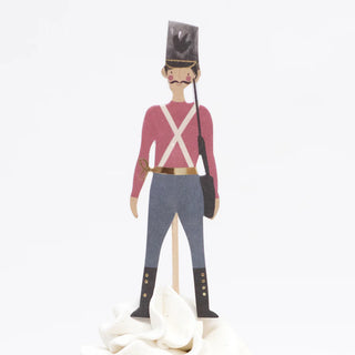 Illustration of a toy soldier from the Meri Meri Nutcracker Cupcake Kit, featuring a tall black hat, pink shirt, blue pants, and boots. He is holding a rifle and standing on a white surface, bringing festive charm to guard your Christmas cupcakes.