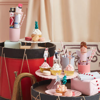 A festive table display showcases Christmas cupcakes in Meri Meri's Nutcracker Cupcake Kit with gold foil cupcake cases, surrounded by macarons and gifts on tiered stands, beautifully adorned with enchanting nutcracker and fairy motifs. Drums and a horn perfectly complement the whimsical arrangement.