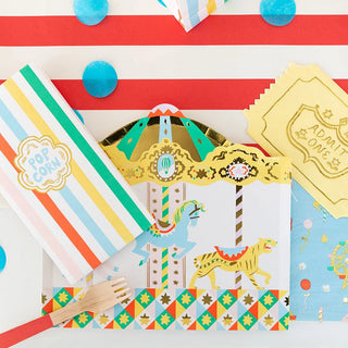 Colorful circus party supplies, including striped popcorn bags, "Off to the Fair" large plates by Daydream Society, a wooden fork, blue confetti, and an "Admit One" ticket, arranged on a red and white striped surface.