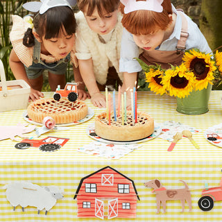 Three children in animal-themed costumes lean over a table with pies and a cake topped with lit candles. The table, adorned with Meri Meri's On The Farm Large Garland and toy tractors, radiates a festive atmosphere. This charming setup features paper cutouts of animals as part of the delightful birthday party decoration.