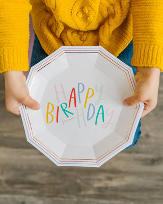 Person holding a Oui Party Hexagon Party Plate from My Mind's Eye with "happy birthday" written in colorful letters, wearing a yellow sweater and blue jeans.