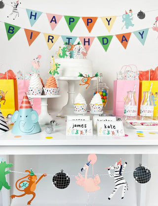 A colorful animal-themed birthday party setup featuring a "Happy Birthday" banner, themed decorations, gift bags, cupcakes, and name cards for James and Kate on a table. Adorning the scene are Merrilulu's Party Animals Garland, fancy drink bottles, party bunting, animal decor, and disco balls to complete the festive look.