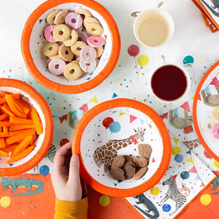 Four "Party Safari Paper Bowls" from the Talking Tables brand hold cookies, biscuits, and sliced carrots on a colorful tablecloth with animal and balloon patterns. Two cups with drinks are also on the table, and a hand is reaching for a bowl at this lively animal-themed party.