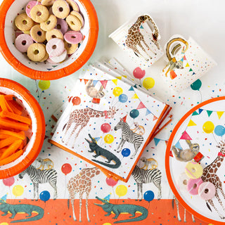 A wildlife-themed party setup featuring jungle-themed partyware like animal illustrations on Talking Tables' Party Safari Paper Napkins and cups, bowls of snacks including cookies and carrot sticks, and a colorful tablecloth and plates adorned with balloons.