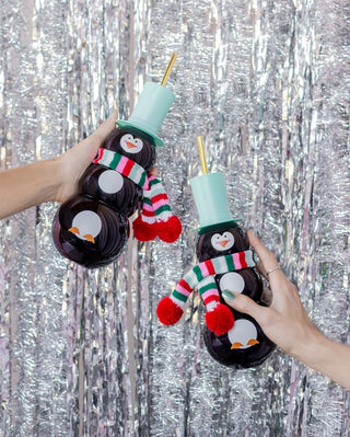 Two people holding a Packed Party Penguin Novelty Sipper in front of a silver background during the holiday season.