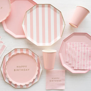 A coordinated set of Petal Pink Signature Cabana Stripe Plates birthday party supplies made with eco-conscious materials from Bonjour Fête, featuring 'happy birthday' text, including plates, cups, and napkins, arranged neatly on.