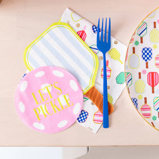 A round pink napkin emblazoned with "LET'S PICKLE" sits on a table set for a Pickleball Social Club gathering, showcasing vibrant Daydream Society Pickleball Social Club Paddle Guest Napkins, accompanied by a blue fork and a patterned plate.