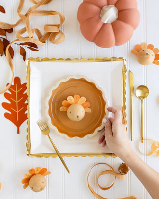 A hand reaches for a plate decorated with a turkey-themed egg, surrounded by autumn-themed elements, golden cutlery, and a pumpkin-shaped decoration—perfect for a Thanksgiving feast or to complement your Pie Dessert Plate collection (My Mind’s Eye).