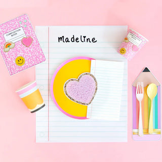 Pink and yellow heart-shaped lunch with Kailo Chic Pink Composition Book Paper Cup, eating utensils, and a notepad with "Madeline" written at the top on a pink background. Perfectly themed for back to school season with all the essential school supplies in place.