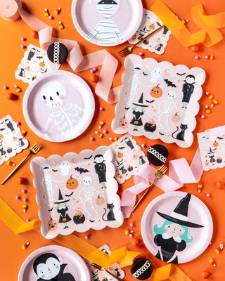 A collection of Halloween-themed paper plates and napkins from the Pink Monster Plate Set by My Mind’s Eye on an orange surface, surrounded by colorful candies, and pink and orange ribbons. Perfect for Halloween parties, the plates feature illustrations of skeletons, witches, and other festive designs.