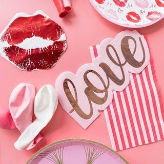 Valentine's themed items on a pink background, showcasing paper lips, a "love" cutout, balloons, and Sprinkle BASH's Pink Striped Guest Napkins—ideal for any pink-themed event.