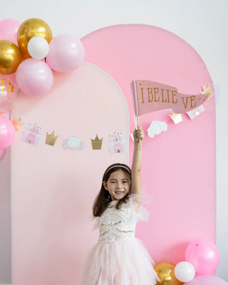 A young girl wearing a pink dress holds a flag that says "I BELIEVE" in front of the My Mind’s Eye Pink Unicorn I Believe Chipboard Pennant Banner and balloon-decorated backdrop at her magical unicorn party.