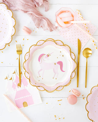 A white table is set with pink and gold unicorn-themed party supplies, including My Mind’s Eye Pink Unicorn Ruffle Edge Plates, napkins, cutlery, macarons, and a beverage in a pink cup with a straw. Confetti is scattered around, creating magical party vibes.