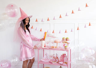 A woman in a pink dress and hat stands beside a whimsically decorated table featuring drinks, treats, and a sign reading "Hocus Pocus." Pink and clear balloons, holiday decorations, and My Mind’s Eye's Pink Witch Hat Banner garland hang as festive decor.