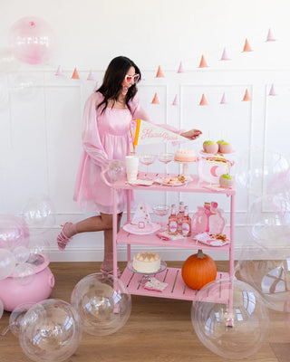 A woman in a pink dress stands next to a decorated cart filled with pink party items, food, and drinks. Clear balloons are scattered on the floor, along with Halloween decor such as a pumpkin and other festive items on the cart. Adding an extra touch of charm to the scene is the Pink Witch Hat Banner from My Mind’s Eye.