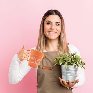 A woman wearing a brown apron holds a small potted plant in one hand and NPW's "Plant Lady Mug" in the other, against a pink background, giving a thumbs-up.