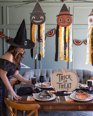 A person dressed as a witch decorates a dining table with Halloween-themed items, including a "Trick or Treat" sign and festive plates, in a room adorned with hanging decorations and garlands. My Mind’s Eye Polka Dot Halloween Dinner Napkins add an extra touch to the spooktacular party decor, making it perfect for a Halloween party.